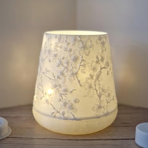 May include: A white, cone-shaped lamp with a floral pattern. The lamp is lit from within, casting a soft glow. The design features delicate white flowers and branches, creating a serene ambiance. The lamp sits on a wooden surface.