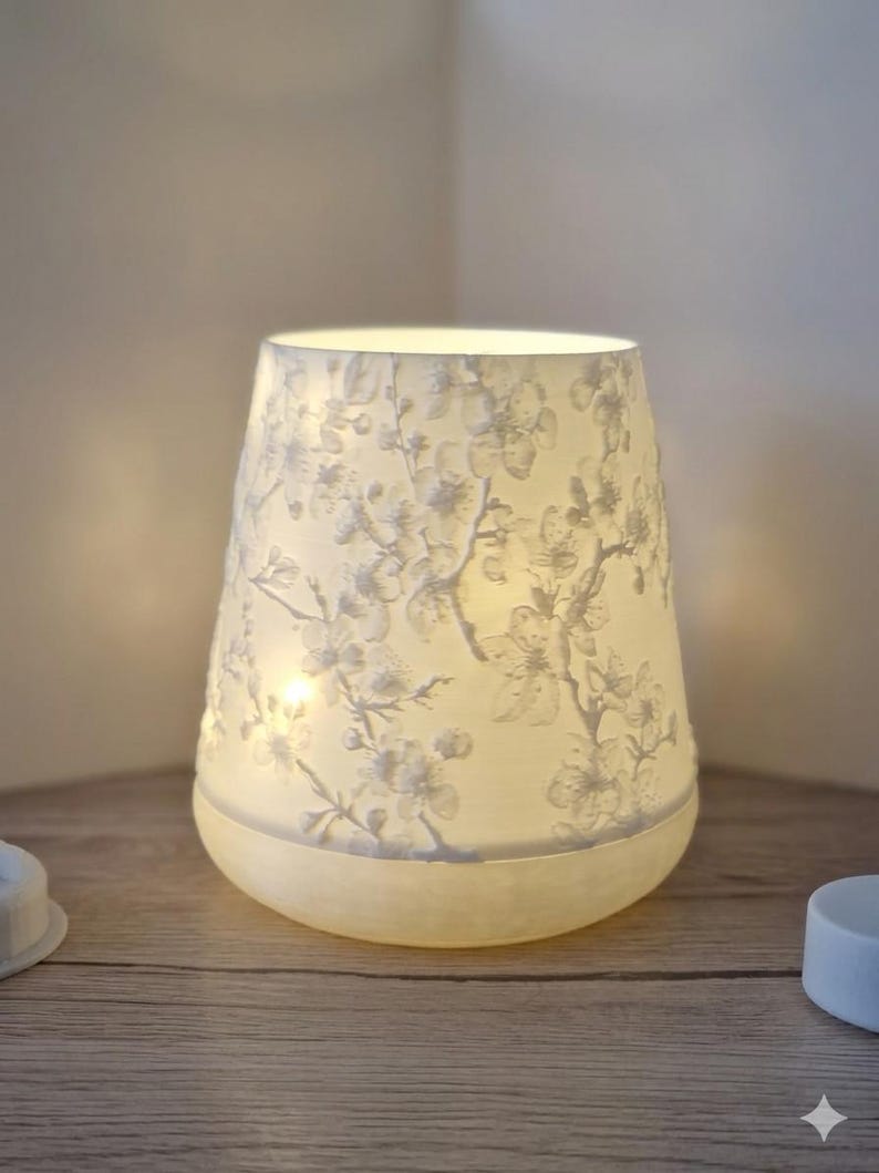 May include: A white, cone-shaped lamp with a floral pattern. The lamp is lit from within, casting a soft glow. The design features delicate white flowers and branches, creating a serene ambiance. The lamp sits on a wooden surface.