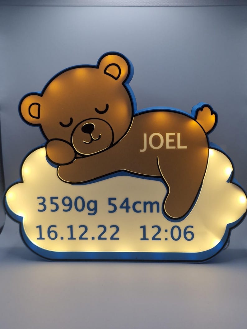 Puede incluir: Una luz de noche con forma de oso de peluche marr&oacute;n con el nombre "JOEL" en una base con forma de nube. La base muestra el peso al nacer del beb&eacute; de 3590g, la longitud de 54cm y la fecha y hora de nacimiento: 16.12.22 12:06.