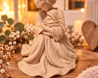 Könnte beinhalten: Eine beige Figur, die eine Person mit hochgestecktem Haar darstellt, die ein strukturiertes Gewand trägt und einen kleinen, in Tücher gewickelten Säugling eng an die Brust hält. Die Skulptur weist detaillierte Schnitzereien an Kleidung und Haar auf.