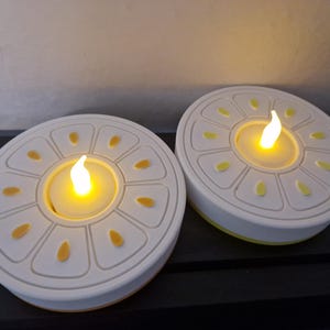 May include: Two white and yellow LED candles shaped like citrus fruit slices. Each candle has a flickering yellow light in the center, resembling a flame. The candles are on a dark surface.