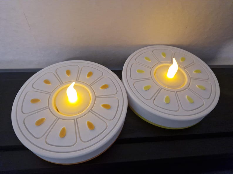 May include: Two white and yellow LED candles shaped like citrus fruit slices. Each candle has a flickering yellow light in the center, resembling a flame. The candles are on a dark surface.