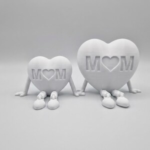 以下が含まれることがあります： 腕と脚が付いた、中央にハートのシンボルが付いた「MOM」という文字が入った、2つの白いハート型の置物。置物は手を繋いでいる。シンプルでモダンなデザインで、家の装飾やギフトに適しています。