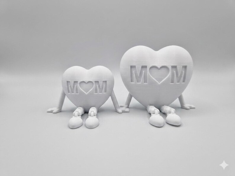 以下が含まれることがあります： 腕と脚が付いた、中央にハートのシンボルが付いた「MOM」という文字が入った、2つの白いハート型の置物。置物は手を繋いでいる。シンプルでモダンなデザインで、家の装飾やギフトに適しています。