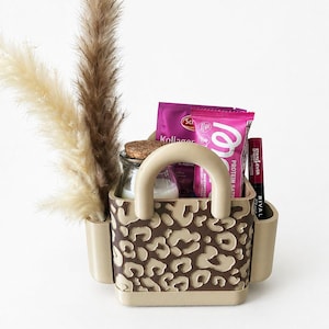 Könnte beinhalten: Eine beige-braune Tragetasche mit Leopardenmuster, die ein kleines Glasgefäß mit Korkdeckel, einen rosa Proteinriegel und eine Tube Lipgloss enthält. Hohes, getrocknetes Ziergras befindet sich im Hintergrund.