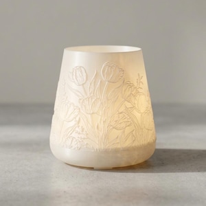 May include: A cream-colored, tulip-decorated candle holder. The ceramic holder has a rounded base and a flared top. The tulip design is embossed around the exterior. The candle holder is lit from within, creating a soft glow.