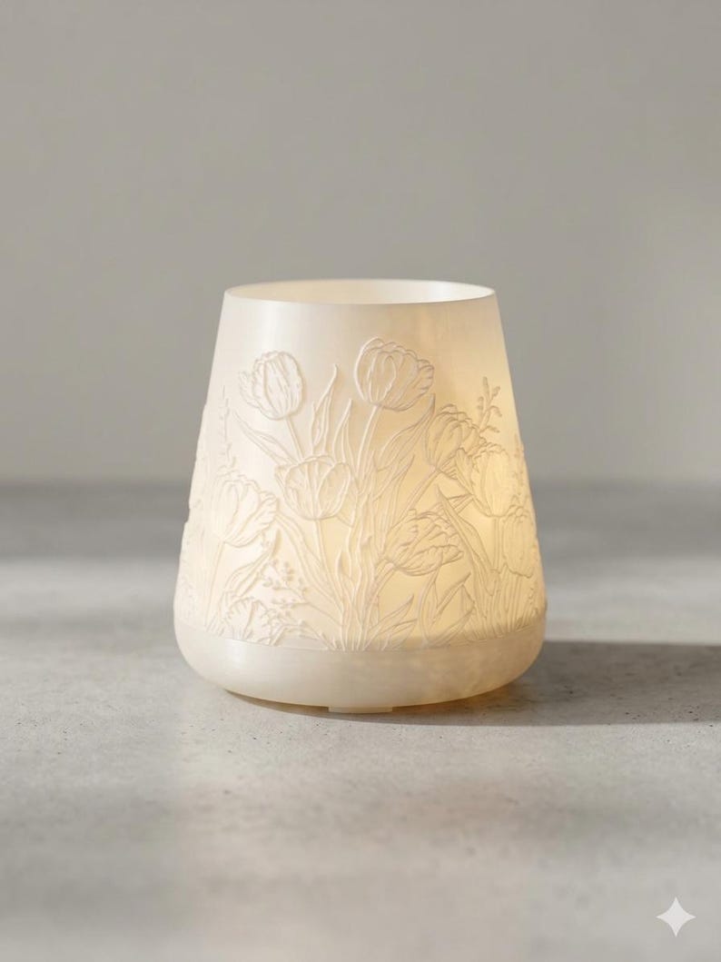 May include: A cream-colored, tulip-decorated candle holder. The ceramic holder has a rounded base and a flared top. The tulip design is embossed around the exterior. The candle holder is lit from within, creating a soft glow.