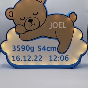 Puede incluir: Una luz nocturna con forma de oso de peluche marr&oacute;n con el nombre "JOEL" en un lado, descansando sobre una base en forma de nube. La base muestra el texto "3590g 54cm 16.12.22 12:06" en azul. La luz tiene un borde azul.