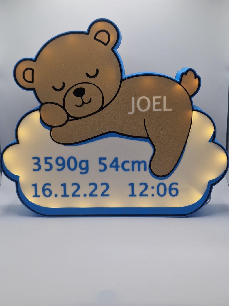 Puede incluir: Una luz nocturna con forma de oso de peluche marr&oacute;n con el nombre "JOEL" en un lado, descansando sobre una base en forma de nube. La base muestra el texto "3590g 54cm 16.12.22 12:06" en azul. La luz tiene un borde azul.