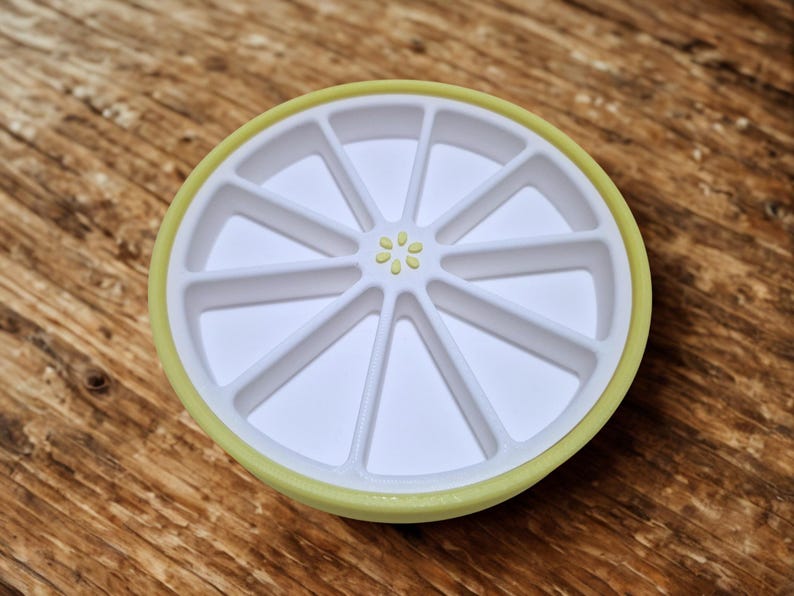 以下が含まれることがあります： 白と黄色のプラスチック製のレモン型トレイで、セグメント化されたコンパートメントがあります。トレイには黄色の縁と中央に小さな花の模様があります。トレイはレモンスライスに似せてデザインされています。