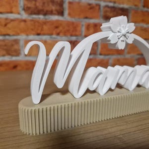 May include: A white 'Mama' sign with a floral detail, set on a beige, ribbed base. The cursive lettering is in a 3D style, with a flower detail at the top of the letter 'M'. The sign is on a wooden surface.