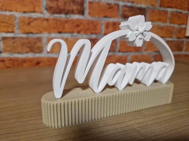 May include: A white 'Mama' sign with a floral detail, set on a beige, ribbed base. The cursive lettering is in a 3D style, with a flower detail at the top of the letter 'M'. The sign is on a wooden surface.