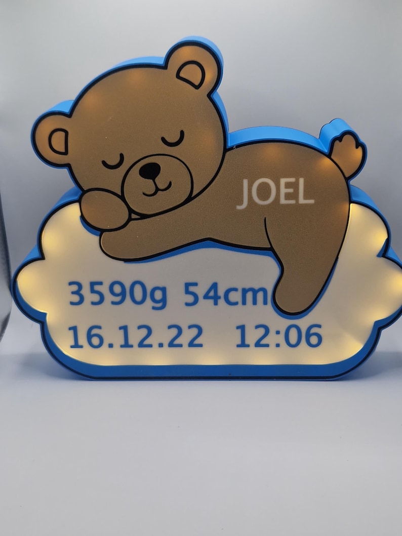 Puede incluir: Una luz de noche con forma de oso de peluche marr&oacute;n con el nombre "JOEL" en un lateral, sobre una base en forma de nube. La base muestra el peso del beb&eacute; (3590g), la longitud (54cm) y la fecha y hora de nacimiento: 16.12.22 12:06. Borde azul.