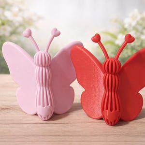 May include: Two butterfly ornaments, one pale pink and one red. Each has heart-shaped antennae and a ribbed body. They are presented on a wooden surface, with a blurred floral backdrop.