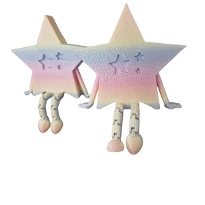 May include: Two whimsical star-shaped figurines with articulated legs and arms. The stars are pastel-colored, with shades of pink, blue, and yellow. Each star has a small star design etched on its surface. The figurines have a playful, cartoonish appearance.