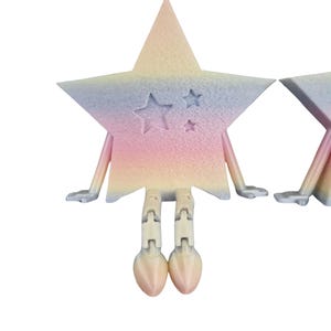 May include: A pastel-colored star-shaped figurine with arms and legs. The star has a gradient of pink, yellow, and blue hues. Three smaller stars are carved into the front. The legs end in rounded feet. The figurine is set against a white background.