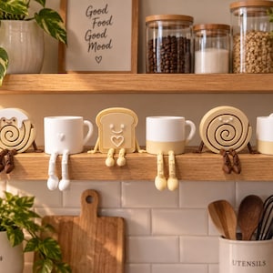 Könnte beinhalten: Eine Sammlung von sechs dekorativen Regalfiguren, die auf einem Holzregal angeordnet sind. Das Set enthält drei Tassen mit baumelnden Beinen und drei lebensmittelthematische Figuren, darunter eine Zimtschnecke, eine Scheibe Toast und ein Keks.