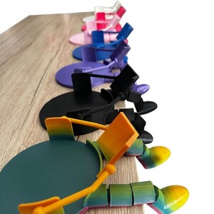 May include: A selection of multi-coloured articulated phone stands. The stands are arranged in a line, displaying a range of hues such as pink, blue, purple, black, and a rainbow gradient. Each stand has a circular base and an adjustable arm.