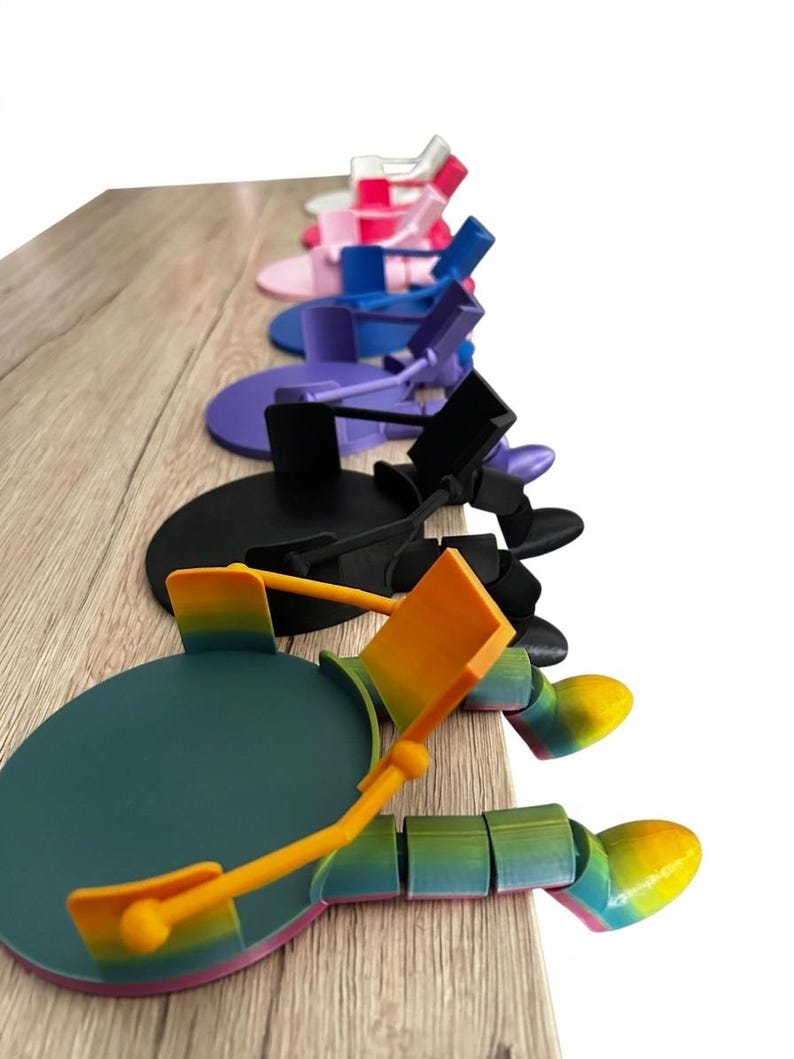 May include: A selection of multi-coloured articulated phone stands. The stands are arranged in a line, displaying a range of hues such as pink, blue, purple, black, and a rainbow gradient. Each stand has a circular base and an adjustable arm.