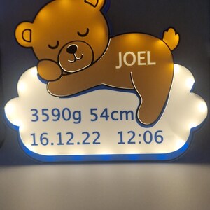 Puede incluir: Un letrero luminoso con un dise&ntilde;o de oso de peluche durmiendo. El oso marr&oacute;n, con un brillo amarillo, descansa sobre una base en forma de nube. El letrero incluye el nombre "JOEL", peso 3590g, altura 54cm, y fecha y hora de nacimiento.