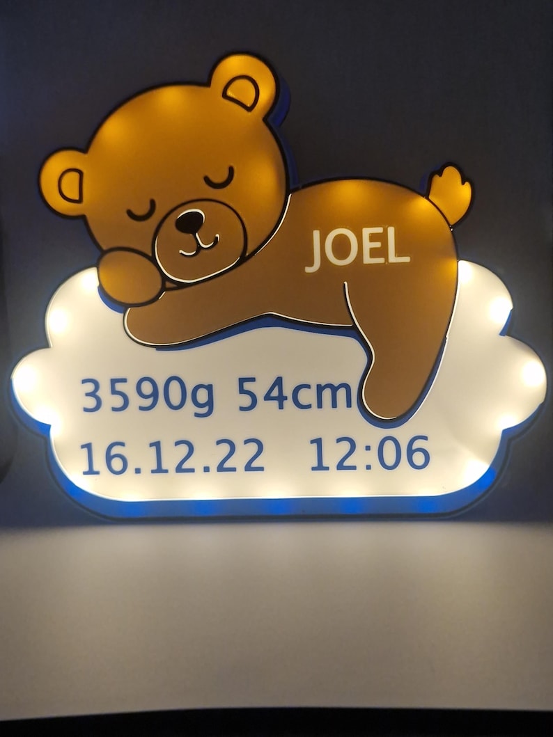 Puede incluir: Un letrero luminoso con un dise&ntilde;o de oso de peluche durmiendo. El oso marr&oacute;n, con un brillo amarillo, descansa sobre una base en forma de nube. El letrero incluye el nombre "JOEL", peso 3590g, altura 54cm, y fecha y hora de nacimiento.