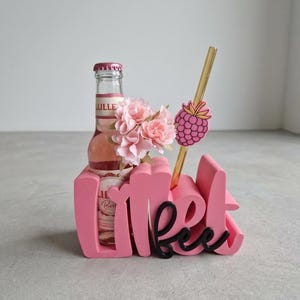 Pode incluir: Um suporte decorativo rosa com a palavra "Lillet" em escrita cursiva. Uma garrafa de Lillet, flores rosas, uma palhinha dourada e uma decora&ccedil;&atilde;o de framboesa est&atilde;o dentro. O suporte est&aacute; numa superf&iacute;cie cinzenta.