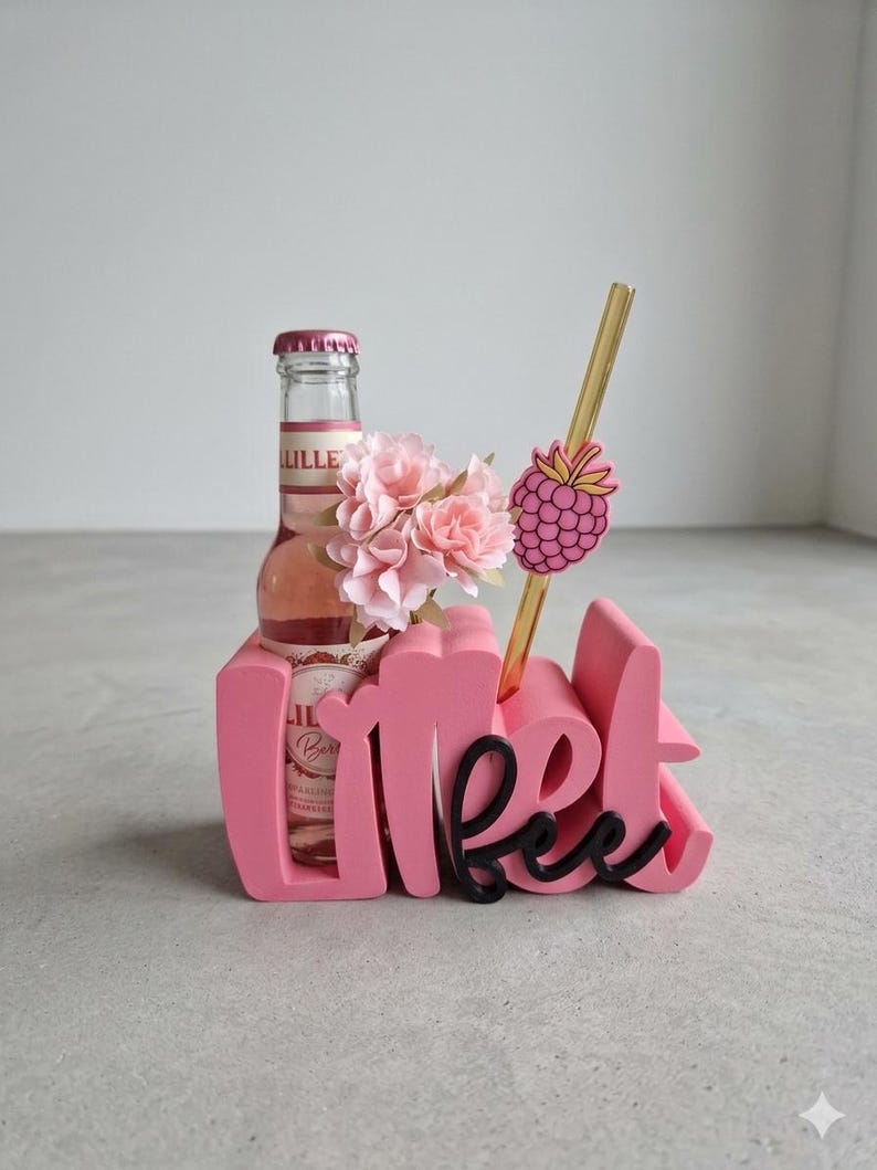 Pode incluir: Um suporte decorativo rosa com a palavra "Lillet" em escrita cursiva. Uma garrafa de Lillet, flores rosas, uma palhinha dourada e uma decora&ccedil;&atilde;o de framboesa est&atilde;o dentro. O suporte est&aacute; numa superf&iacute;cie cinzenta.