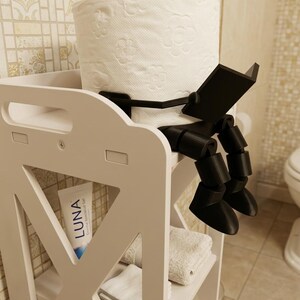 May include: A white bathroom storage unit with a roll of toilet paper held by a black, reading-figure-shaped holder. The unit has shelves with towels and a tube of Luna gel.