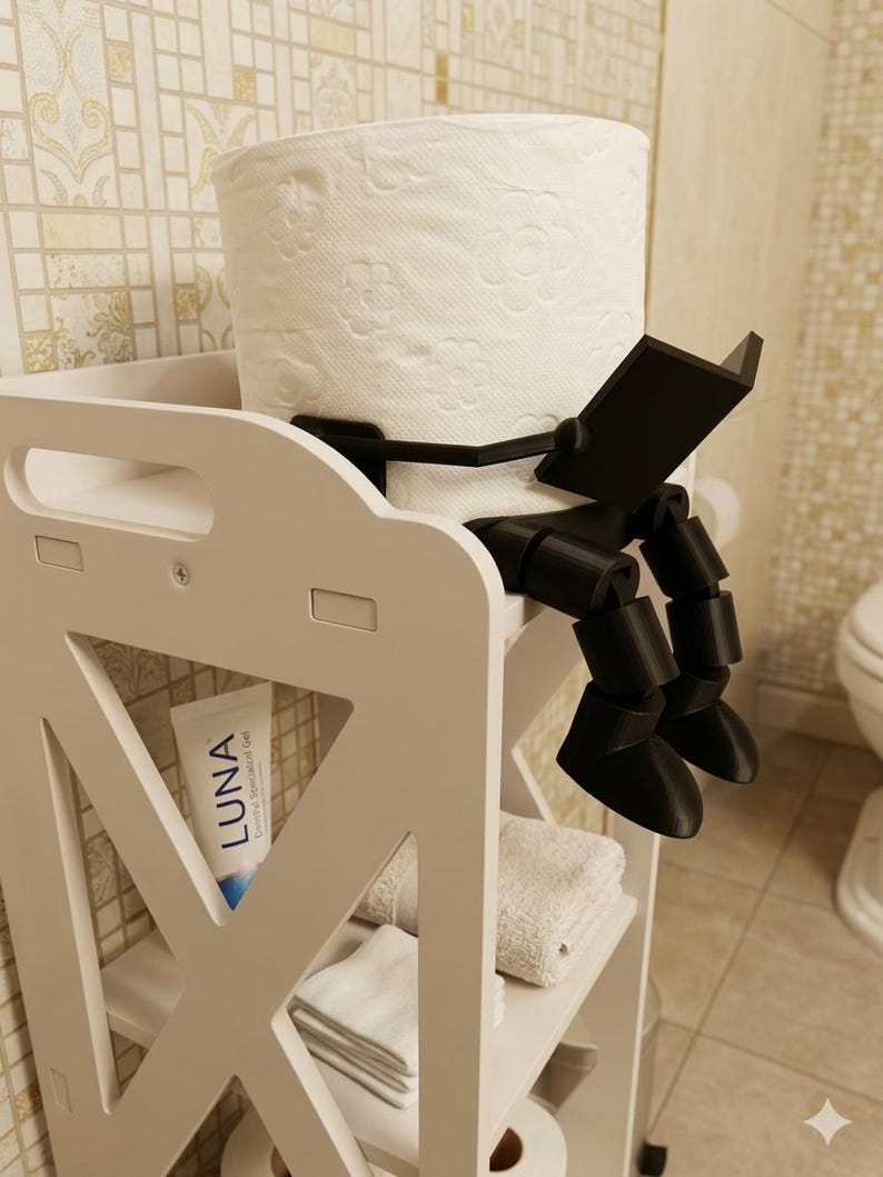 May include: A white bathroom storage unit with a roll of toilet paper held by a black, reading-figure-shaped holder. The unit has shelves with towels and a tube of Luna gel.
