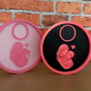 Op de afbeelding: Twee ronde decoratieve items met een moeder-en-kind-ontwerp. E&eacute;n is lichtroze, de andere zwart, beide met een roze rand. De moeder en het kind omarmen elkaar liefdevol, met een hartje erboven. De achtergrond is een bakstenen muur.
