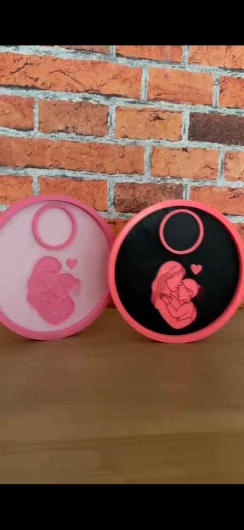 Op de afbeelding: Twee ronde decoratieve items met een moeder-en-kind-ontwerp. E&eacute;n is lichtroze, de andere zwart, beide met een roze rand. De moeder en het kind omarmen elkaar liefdevol, met een hartje erboven. De achtergrond is een bakstenen muur.
