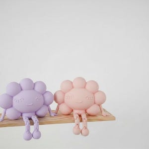 K&ouml;nnte beinhalten: Zwei skurrile, bl&uuml;tenf&ouml;rmige Figuren in Pastellfarben, eine in Lavendel und eine in Pfirsich, sitzen auf einem kleinen Holzregal. Jede Figur hat ein l&auml;chelndes Gesicht, Arme und h&auml;ngende Beine. Der Hintergrund ist schlicht wei&szlig;.