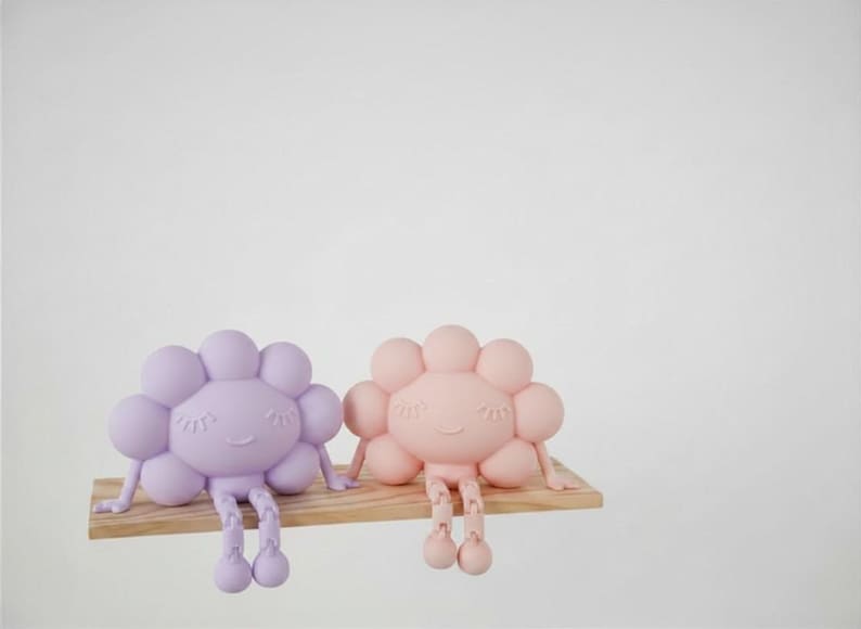 K&ouml;nnte beinhalten: Zwei skurrile, bl&uuml;tenf&ouml;rmige Figuren in Pastellfarben, eine in Lavendel und eine in Pfirsich, sitzen auf einem kleinen Holzregal. Jede Figur hat ein l&auml;chelndes Gesicht, Arme und h&auml;ngende Beine. Der Hintergrund ist schlicht wei&szlig;.