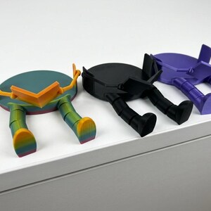 May include: Three quirky, colourful, 3D-printed figures with circular bodies and legs. One is teal with orange accents and rainbow legs, another is black, and the third is purple. They appear to be resting on a white surface.