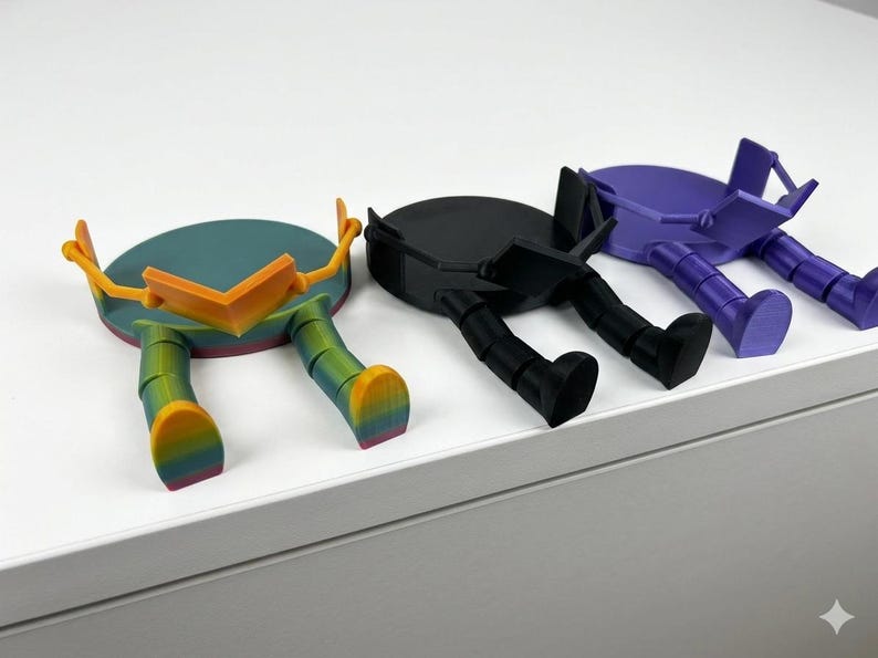 May include: Three quirky, colourful, 3D-printed figures with circular bodies and legs. One is teal with orange accents and rainbow legs, another is black, and the third is purple. They appear to be resting on a white surface.