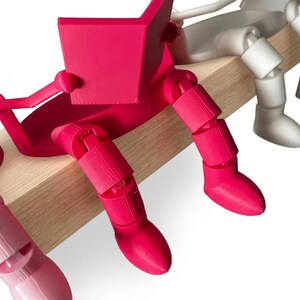 May include: A row of articulated figures in various colours, sitting on a wooden beam. The figures are reading books. The first figure is bright pink, the second is light pink, and the third is white. The figures have segmented limbs.