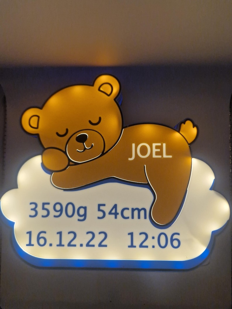 Puede incluir: Ilustraci&oacute;n de un oso de peluche amarillo dorado con el nombre "JOEL" en un letrero blanco con forma de nube. El letrero muestra los detalles del nacimiento del beb&eacute;: 3590g, 54cm, y la fecha y hora de nacimiento: 16.12.22 12:06.