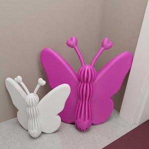 May include: Two decorative butterfly wall art pieces. One is white, the other is bright pink. Both have heart-shaped antennae and a ribbed body. The butterflies are made of a solid material and are placed against a neutral-coloured wall.