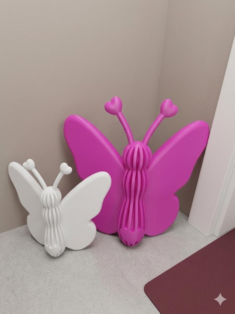 May include: Two decorative butterfly wall art pieces. One is white, the other is bright pink. Both have heart-shaped antennae and a ribbed body. The butterflies are made of a solid material and are placed against a neutral-coloured wall.