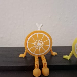 May include: A whimsical orange slice figurine with articulated legs and arms. The orange slice is yellow with white segments and a white stem. The figurine is standing on a dark surface.