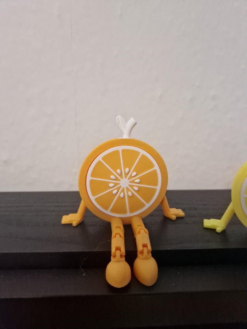 May include: A whimsical orange slice figurine with articulated legs and arms. The orange slice is yellow with white segments and a white stem. The figurine is standing on a dark surface.