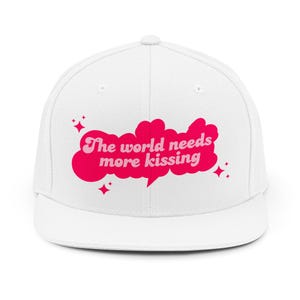 Op de afbeelding: Witte baseballcap met platte klep, met de roze tekst "The world needs more kissing". De tekst staat in een tekstballon, met roze steraccenten.
