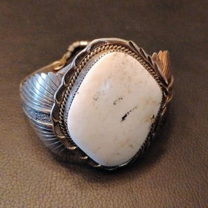 May include: A silver cuff bracelet with a large, off-white, rectangular stone as the centerpiece. The stone is framed by a decorative silver setting with a rope-like border. The bracelet has a detailed, feather-like design on the sides.