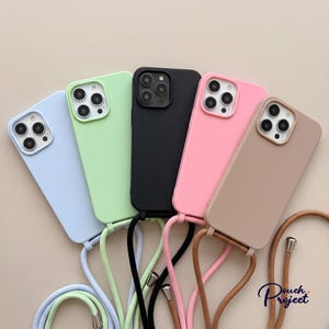 May include: Five smartphone cases with attached crossbody lanyards arranged in a fan shape. The cases are light blue, light green, black, pink, and beige. Each case features a square camera cutout and a matching colored cord with metallic end caps.