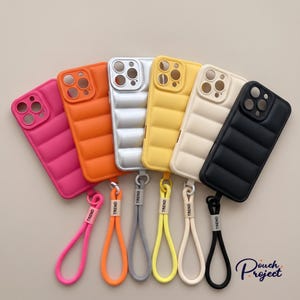 May include: A collection of six puffer-style phone cases in pink, orange, silver, yellow, cream, and black. Each case features a quilted design, a camera cutout, and a matching wrist strap with a small tag labeled TREND attached to the bottom.
