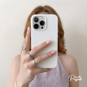 May include: A white phone case with a camera module is held in front of a person. The person's hand, with rings and pink nail polish, is visible. The phone case has a matte finish. The background is a soft, neutral colour.