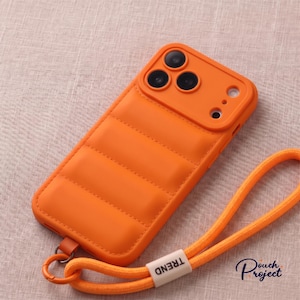 Könnte beinhalten: Eine leuchtend orangefarbene Smartphone-Hülle mit einer gesteppten Puffer-Textur auf der Rückseite. Die Hülle verfügt über einen erhöhten Kameraausschnitt und ein passendes orangefarbenes gewebtes Trageband mit einem weißen Etikett, das TREND zeigt.