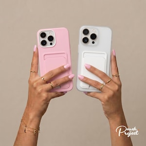 Puede incluir: Dos fundas para smartphone, una en rosa claro sólido y otra en blanco translúcido, cada una con un bolsillo rectangular para tarjetas en la parte posterior. Las fundas tienen un recorte para triple cámara y se sostienen frente a un fondo neutro.