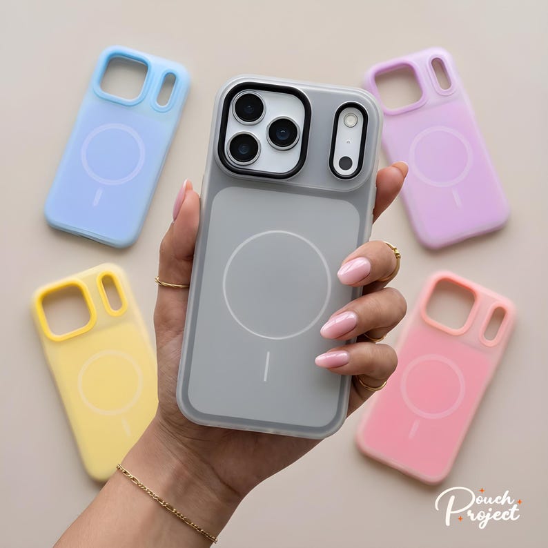 Pu&ograve; includere: Una mano che tiene una custodia grigia per telefono con un bordo trasparente, circondata da custodie colorate in blu, giallo, rosa e viola. Ogni custodia ha un design circolare e un simbolo di ricarica magnetica.