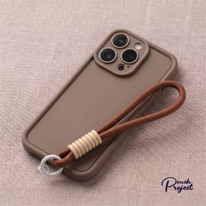 Op de afbeelding: Een matbruine smartphonehoes met een uitsparing voor een drievoudige camera en een bruine polsband van koord. De band is bevestigd met een witte plastic ring en geribbelde, crèmekleurige siliconen kralen. De tekst Pouch Project is zichtbaar.
